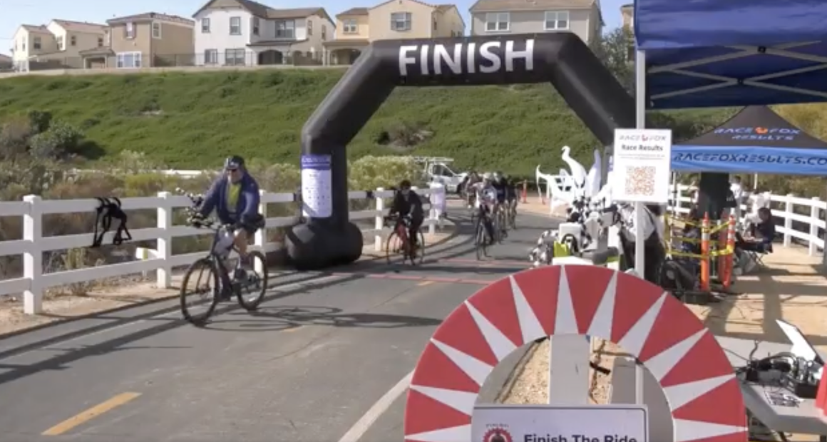 Alt text: Cyclists cross the finish line at an outdoor bike race on a paved path lined with white fences. A large black inflatable arch with the word “FINISH” marks the end of the course, with several riders approaching. On the right, a blue tent labeled “RaceFox Results” houses event staff and race equipment. Houses and green hills are visible in the background under clear skies.