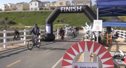 Alt text: Cyclists cross the finish line at an outdoor bike race on a paved path lined with white fences. A large black inflatable arch with the word “FINISH” marks the end of the course, with several riders approaching. On the right, a blue tent labeled “RaceFox Results” houses event staff and race equipment. Houses and green hills are visible in the background under clear skies.