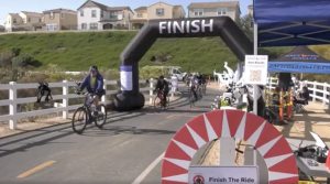 Alt text: Cyclists cross the finish line at an outdoor bike race on a paved path lined with white fences. A large black inflatable arch with the word “FINISH” marks the end of the course, with several riders approaching. On the right, a blue tent labeled “RaceFox Results” houses event staff and race equipment. Houses and green hills are visible in the background under clear skies.