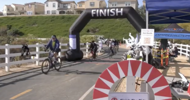 Alt text: Cyclists cross the finish line at an outdoor bike race on a paved path lined with white fences. A large black inflatable arch with the word “FINISH” marks the end of the course, with several riders approaching. On the right, a blue tent labeled “RaceFox Results” houses event staff and race equipment. Houses and green hills are visible in the background under clear skies.
