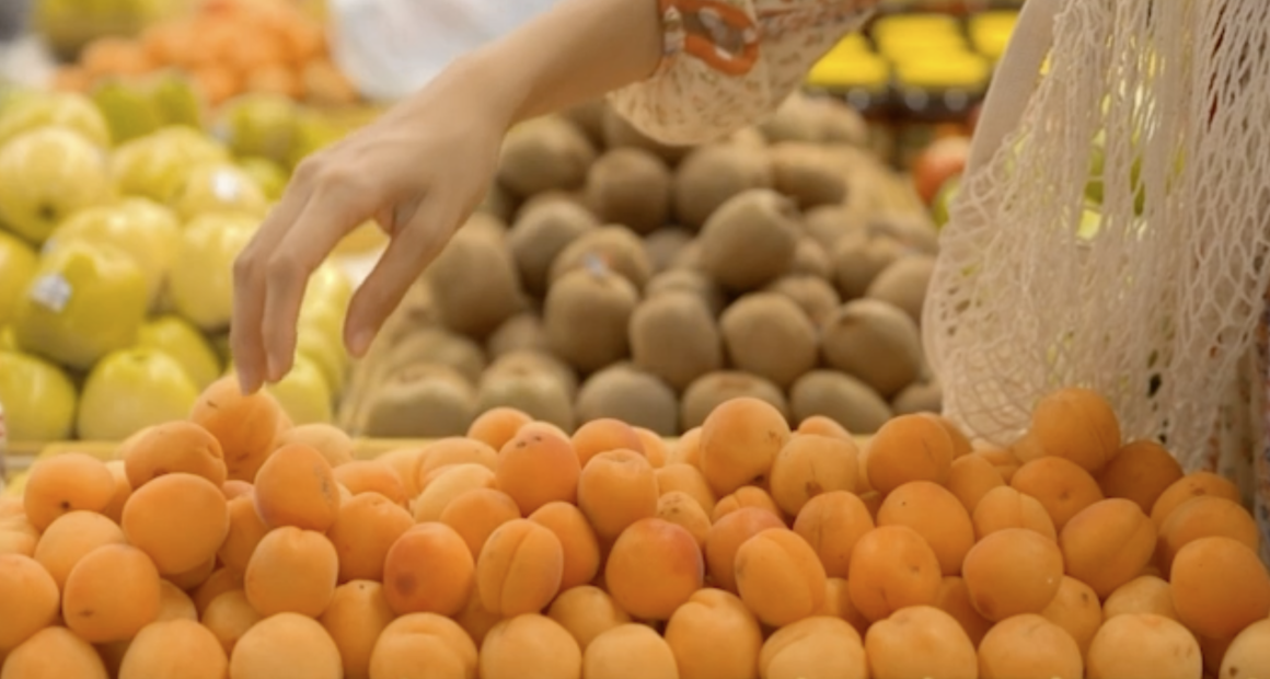 A person’s hand reaches out to pick an apricot from a display of fresh apricots in a grocery store. Other fruits, including kiwis and apples, are visible in the background, and a reusable mesh bag hangs from the person’s arm.