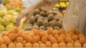A person’s hand reaches out to pick an apricot from a display of fresh apricots in a grocery store. Other fruits, including kiwis and apples, are visible in the background, and a reusable mesh bag hangs from the person’s arm.