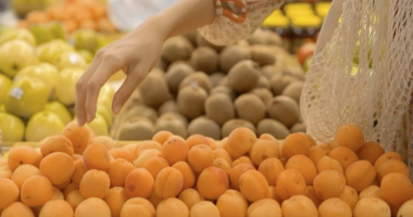 A person’s hand reaches out to pick an apricot from a display of fresh apricots in a grocery store. Other fruits, including kiwis and apples, are visible in the background, and a reusable mesh bag hangs from the person’s arm.
