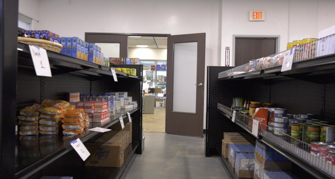 Shelves stocked with nonperishable food items such as canned goods, boxed pasta, and instant noodles line both sides of an aisle in a small pantry or food bank. Signs on the shelves indicate item limits, and an open door at the end of the aisle reveals another room with additional supplies.