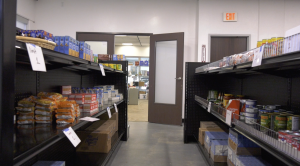 Shelves stocked with nonperishable food items such as canned goods, boxed pasta, and instant noodles line both sides of an aisle in a small pantry or food bank. Signs on the shelves indicate item limits, and an open door at the end of the aisle reveals another room with additional supplies.