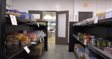 Shelves stocked with nonperishable food items such as canned goods, boxed pasta, and instant noodles line both sides of an aisle in a small pantry or food bank. Signs on the shelves indicate item limits, and an open door at the end of the aisle reveals another room with additional supplies.