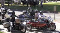 A group of motorcyclists, some riding with sidecars, are parked or slowly riding on a paved road lined with grass and trees. The closest motorcyclist on the left is on a gray touring bike. To the right, a couple is riding a red and black motorcycle with a sidecar. In the background, people on horses are visible.