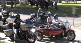 A group of motorcyclists, some riding with sidecars, are parked or slowly riding on a paved road lined with grass and trees. The closest motorcyclist on the left is on a gray touring bike. To the right, a couple is riding a red and black motorcycle with a sidecar. In the background, people on horses are visible.