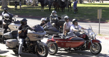 A group of motorcyclists, some riding with sidecars, are parked or slowly riding on a paved road lined with grass and trees. The closest motorcyclist on the left is on a gray touring bike. To the right, a couple is riding a red and black motorcycle with a sidecar. In the background, people on horses are visible.