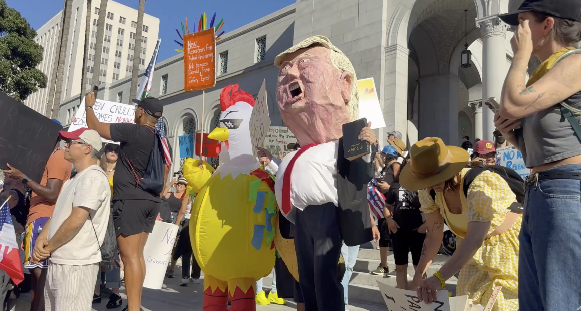 A crowd of protesters gathers outside a large government building, holding various signs and banners. Among them are people wearing costumes, including one person in a yellow chicken suit and another with a large caricature mask resembling a political figure. The scene is bright and sunny, with participants displaying messages related to political and social issues.
