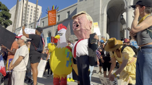 A crowd of protesters gathers outside a large government building, holding various signs and banners. Among them are people wearing costumes, including one person in a yellow chicken suit and another with a large caricature mask resembling a political figure. The scene is bright and sunny, with participants displaying messages related to political and social issues.