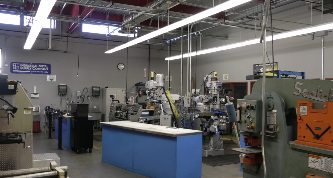 A metalworking shop with various industrial machines, including mills, lathes, and presses. The workspace has gray walls, overhead fluorescent lighting, exposed ceiling ducts, and a blue workbench in the center. A sign reading “Industrial Metal Supply Company” hangs on the wall.
