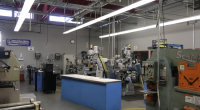 A metalworking shop with various industrial machines, including mills, lathes, and presses. The workspace has gray walls, overhead fluorescent lighting, exposed ceiling ducts, and a blue workbench in the center. A sign reading “Industrial Metal Supply Company” hangs on the wall.