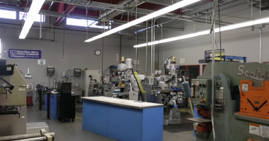 A metalworking shop with various industrial machines, including mills, lathes, and presses. The workspace has gray walls, overhead fluorescent lighting, exposed ceiling ducts, and a blue workbench in the center. A sign reading “Industrial Metal Supply Company” hangs on the wall.