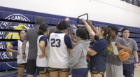 A women's basketball team and their coach are huddled together in a gym, forming a tight circle with their hands reaching toward the center. Several players are wearing white and navy blue jerseys with gold trim, and one player's jersey shows the number 23. The background features blue bleachers with a large yellow and black mascot logo and team name. One player on the far right holds a Wilson basketball and looks toward the camera.