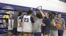 A women's basketball team and their coach are huddled together in a gym, forming a tight circle with their hands reaching toward the center. Several players are wearing white and navy blue jerseys with gold trim, and one player's jersey shows the number 23. The background features blue bleachers with a large yellow and black mascot logo and team name. One player on the far right holds a Wilson basketball and looks toward the camera.