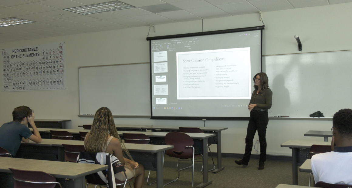 A woman stands at the front of a classroom giving a presentation on obsessive-compulsive disorder (OCD) to a small group of students. A slide projected behind her lists “Some Common Compulsions.” Students sit at desks listening attentively, and a large periodic table poster hangs on the wall to the left.