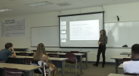 A woman stands at the front of a classroom giving a presentation on obsessive-compulsive disorder (OCD) to a small group of students. A slide projected behind her lists “Some Common Compulsions.” Students sit at desks listening attentively, and a large periodic table poster hangs on the wall to the left.