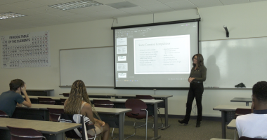 A woman stands at the front of a classroom giving a presentation on obsessive-compulsive disorder (OCD) to a small group of students. A slide projected behind her lists “Some Common Compulsions.” Students sit at desks listening attentively, and a large periodic table poster hangs on the wall to the left.