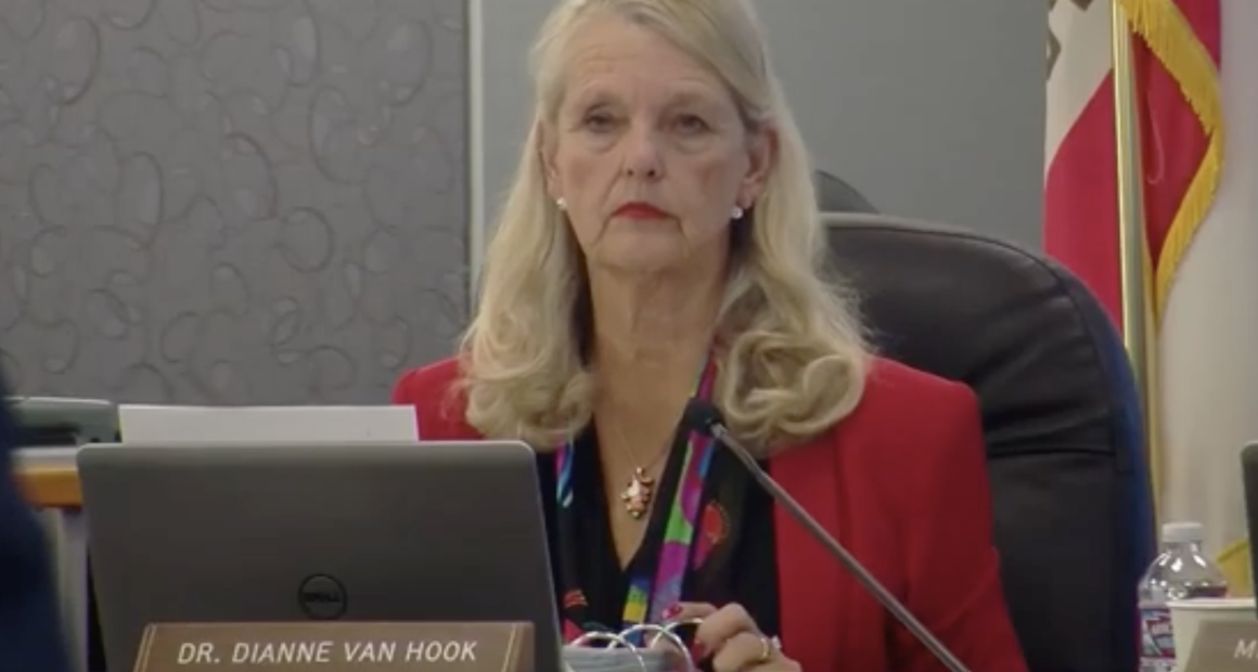 Dr. Dianne Van Hook, sitting at a large dark table during a meeting. She is looking directly at the camera with a neutral, serious expression. She is wearing a bright red blazer over a black top with a multi-colored floral or patterned scarf/lanyard. A laptop is open in front of her. To the right, a partially visible flag (likely the California state flag) and a bottle of water are present.