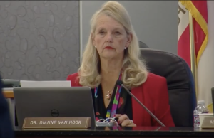 Dr. Dianne Van Hook, sitting at a large dark table during a meeting. She is looking directly at the camera with a neutral, serious expression. She is wearing a bright red blazer over a black top with a multi-colored floral or patterned scarf/lanyard. A laptop is open in front of her. To the right, a partially visible flag (likely the California state flag) and a bottle of water are present.