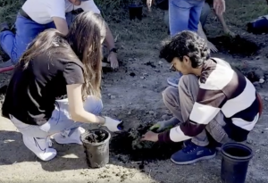 a COC student in black on the left helps another student in a brown and white striped shirt dig a hole in the ground for a small plant.