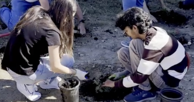 a COC student in black on the left helps another student in a brown and white striped shirt dig a hole in the ground for a small plant.