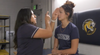 An athletic trainer performs an eye-movement test on a student athlete in a training room. The trainer holds up one hand and points while the athlete, sitting on a treatment table with one eye covered, focuses straight ahead. Both are wearing navy College of the Canyons shirts, and a wall pad with the school’s sports medicine logo is visible behind them