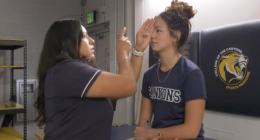 An athletic trainer performs an eye-movement test on a student athlete in a training room. The trainer holds up one hand and points while the athlete, sitting on a treatment table with one eye covered, focuses straight ahead. Both are wearing navy College of the Canyons shirts, and a wall pad with the school’s sports medicine logo is visible behind them