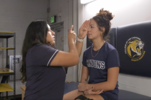 An athletic trainer performs an eye-movement test on a student athlete in a training room. The trainer holds up one hand and points while the athlete, sitting on a treatment table with one eye covered, focuses straight ahead. Both are wearing navy College of the Canyons shirts, and a wall pad with the school’s sports medicine logo is visible behind them
