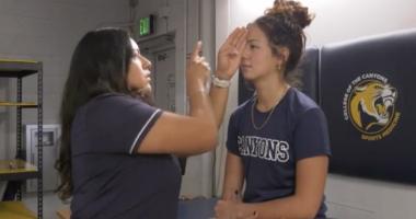 An athletic trainer performs an eye-movement test on a student athlete in a training room. The trainer holds up one hand and points while the athlete, sitting on a treatment table with one eye covered, focuses straight ahead. Both are wearing navy College of the Canyons shirts, and a wall pad with the school’s sports medicine logo is visible behind them