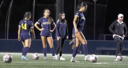 Four women on a soccer field at night prepare for practice or warm-ups. Three players wear navy uniforms with “Canyons” across the front, and one wears a navy warm-up jacket. Two soccer balls sit on the turf in front of them. One player on the right steps toward a ball, while the others stand nearby, watching or waiting. Stadium lights illuminate the scene against a dark background.