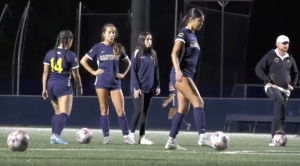 Four women on a soccer field at night prepare for practice or warm-ups. Three players wear navy uniforms with “Canyons” across the front, and one wears a navy warm-up jacket. Two soccer balls sit on the turf in front of them. One player on the right steps toward a ball, while the others stand nearby, watching or waiting. Stadium lights illuminate the scene against a dark background.