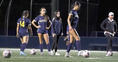 Four women on a soccer field at night prepare for practice or warm-ups. Three players wear navy uniforms with “Canyons” across the front, and one wears a navy warm-up jacket. Two soccer balls sit on the turf in front of them. One player on the right steps toward a ball, while the others stand nearby, watching or waiting. Stadium lights illuminate the scene against a dark background.