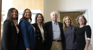 A group of six adults stand together indoors, smiling at the camera. They are dressed in professional attire: five women wearing business or chef-style clothing and one man in a blazer and button-down shirt. They are standing in a line, posing for a group photo in front of a light-colored wall and a sign partially reading “Cul… Arts.”