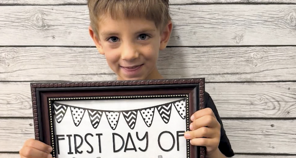 A young child smiles while holding a framed sign that reads “First Day of Kindergarten.” The background is a light wood plank wall.