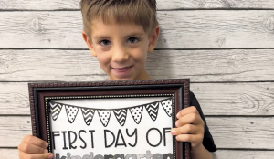 A young child smiles while holding a framed sign that reads “First Day of Kindergarten.” The background is a light wood plank wall.