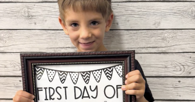 A young child smiles while holding a framed sign that reads “First Day of Kindergarten.” The background is a light wood plank wall.