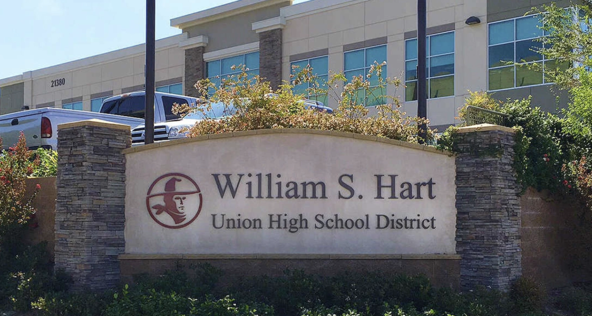 Here’s an appropriate alt text description for the image: **Alt text:** *A stone monument sign reading “William S. Hart Union High School District” stands in front of a modern two-story building with large windows. The sign features a red logo of a frontier-style figure in profile. Landscaping with bushes and flowers surrounds the sign, and several parked trucks are visible behind it.*