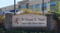 Here’s an appropriate alt text description for the image: **Alt text:** *A stone monument sign reading “William S. Hart Union High School District” stands in front of a modern two-story building with large windows. The sign features a red logo of a frontier-style figure in profile. Landscaping with bushes and flowers surrounds the sign, and several parked trucks are visible behind it.*