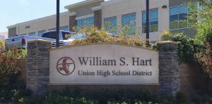 Here’s an appropriate alt text description for the image: **Alt text:** *A stone monument sign reading “William S. Hart Union High School District” stands in front of a modern two-story building with large windows. The sign features a red logo of a frontier-style figure in profile. Landscaping with bushes and flowers surrounds the sign, and several parked trucks are visible behind it.*