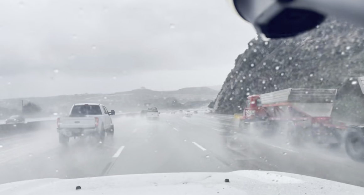 A rainy highway scene viewed through a wet windshield, with several vehicles driving through heavy spray. A white pickup truck is ahead in the left lane, and a large red truck with a trailer is passing on the right. Low hills and an overcast sky are visible in the background.