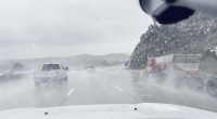 A rainy highway scene viewed through a wet windshield, with several vehicles driving through heavy spray. A white pickup truck is ahead in the left lane, and a large red truck with a trailer is passing on the right. Low hills and an overcast sky are visible in the background.