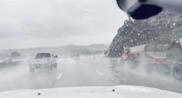 A rainy highway scene viewed through a wet windshield, with several vehicles driving through heavy spray. A white pickup truck is ahead in the left lane, and a large red truck with a trailer is passing on the right. Low hills and an overcast sky are visible in the background.
