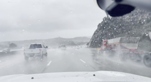A rainy highway scene viewed through a wet windshield, with several vehicles driving through heavy spray. A white pickup truck is ahead in the left lane, and a large red truck with a trailer is passing on the right. Low hills and an overcast sky are visible in the background.