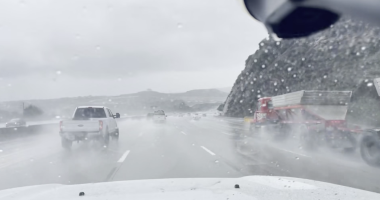A rainy highway scene viewed through a wet windshield, with several vehicles driving through heavy spray. A white pickup truck is ahead in the left lane, and a large red truck with a trailer is passing on the right. Low hills and an overcast sky are visible in the background.