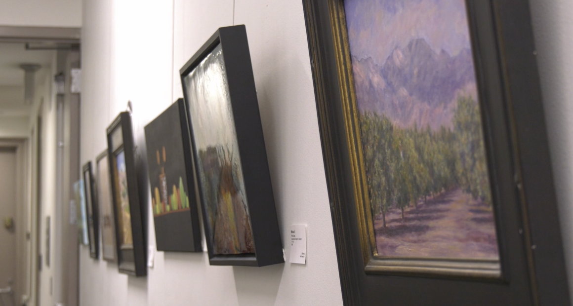 A row of framed paintings hung on a bright white wall in an art gallery or hallway. The paintings, which appear to be mostly landscapes and abstract pieces, are framed in black or dark wood and recede into the distance on the left. The painting closest to the viewer on the right depicts a green field or orchard leading up to purple mountains under a hazy sky.