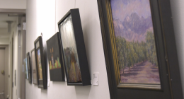 A row of framed paintings hung on a bright white wall in an art gallery or hallway. The paintings, which appear to be mostly landscapes and abstract pieces, are framed in black or dark wood and recede into the distance on the left. The painting closest to the viewer on the right depicts a green field or orchard leading up to purple mountains under a hazy sky.