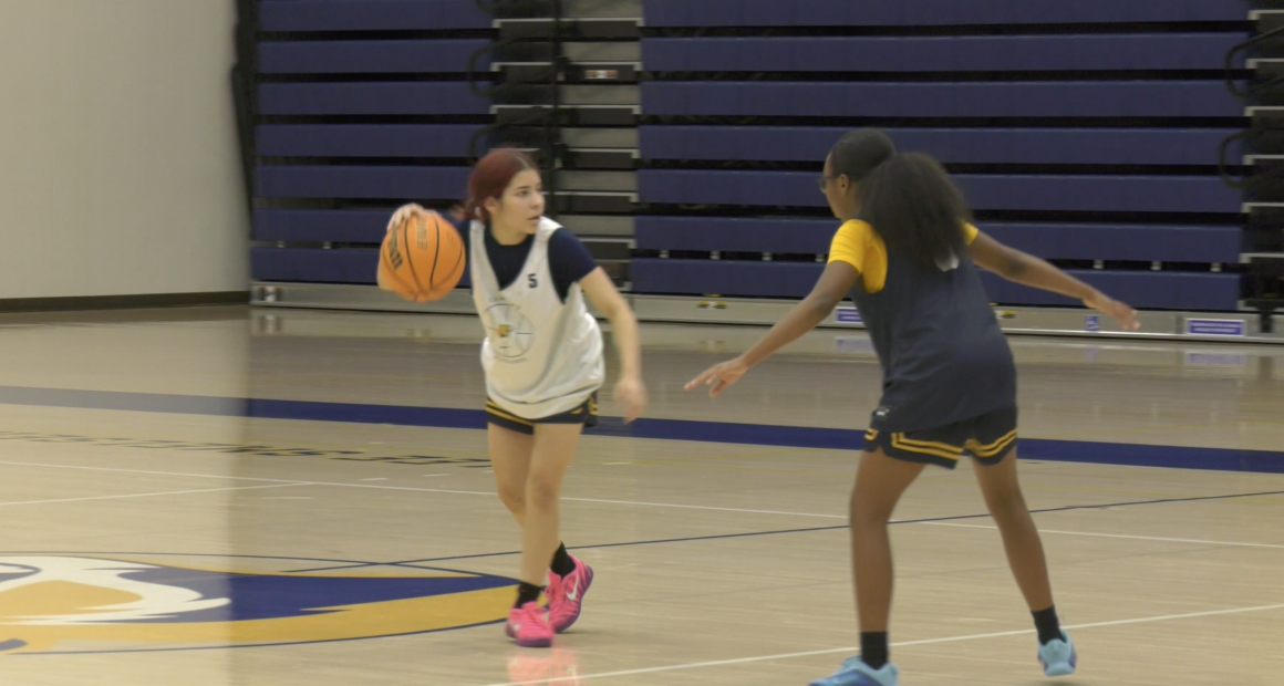 Two basketball players scrimmage on an indoor court. One player in a white practice jersey dribbles the ball while being guarded closely by another player in a dark practice jersey with yellow sleeves. The gym’s bleachers are visible in the background.