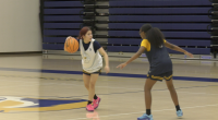 Two basketball players scrimmage on an indoor court. One player in a white practice jersey dribbles the ball while being guarded closely by another player in a dark practice jersey with yellow sleeves. The gym’s bleachers are visible in the background.